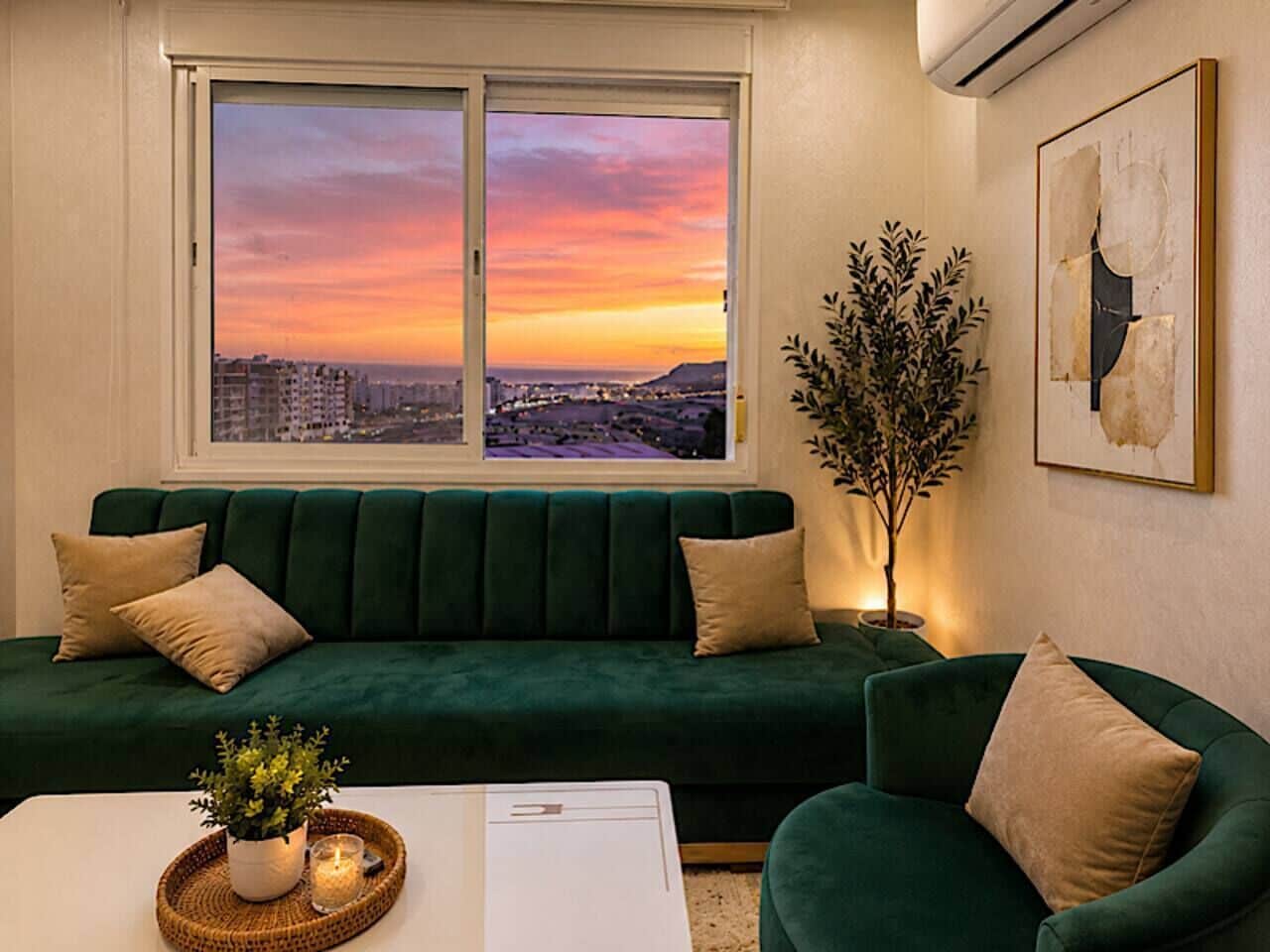 A green sofa with two pillows, a small table with a plant, and a window with a cityscape view.