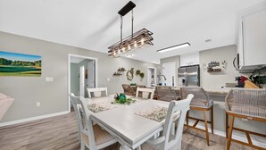 A dining area with a white table and chairs, a hanging light fixture, and a kitchen with white cabinets. - First Floor condo on True Blue Golf course with Pool!!! (Pawleys Island)