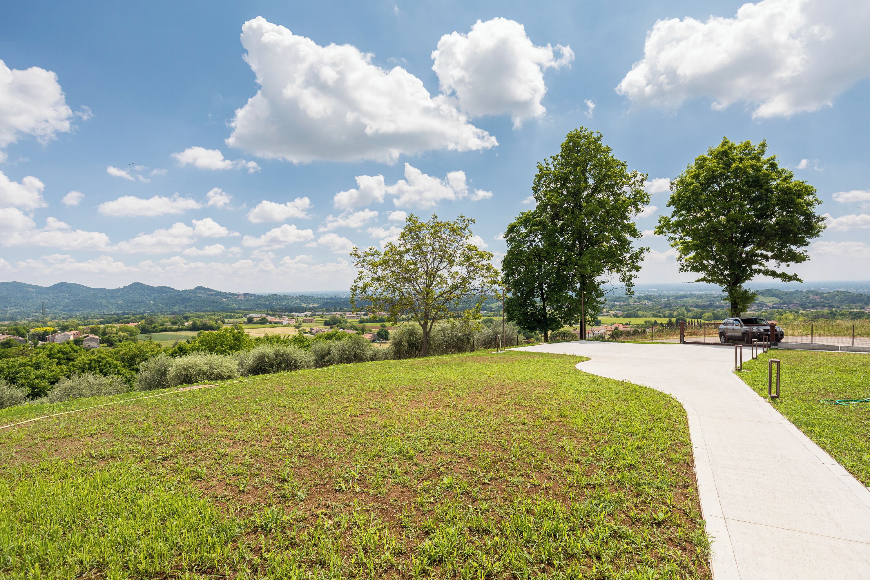 A paved pathway curving through a grassy area with trees and a distant view of hills.