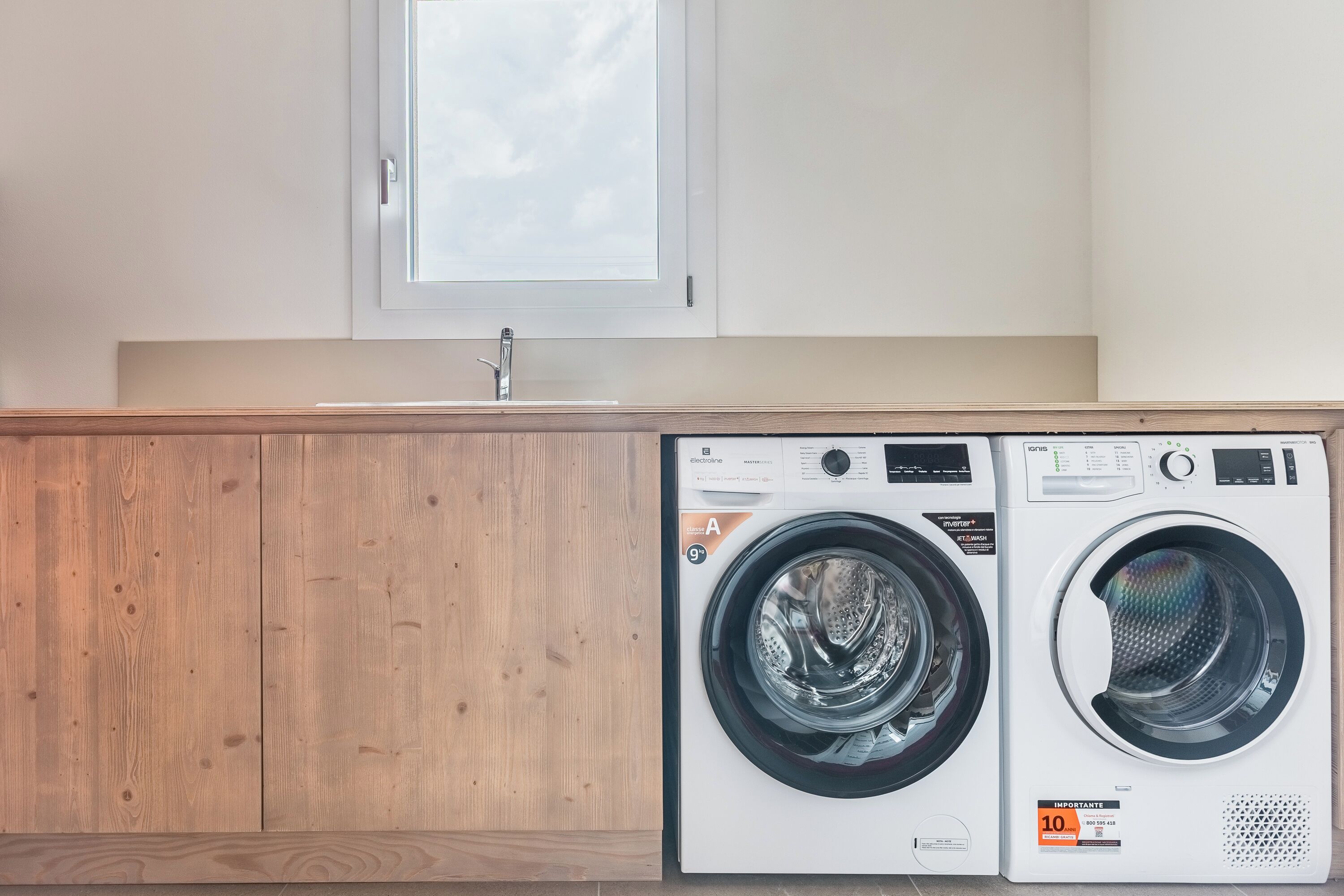 A laundry area with two front-loading washing machines, a wooden cabinet, and a window above the sink.