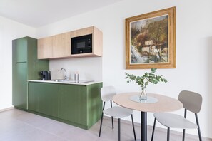 Eine moderne Küche mit grünen Schränken, einer Mikrowelle und einer Kaffeemaschine. Ein Esstisch mit Stühlen und eine Pflanze in einer Vase stehen dort. Ein gerahmtes Gemälde ist an der Wand.