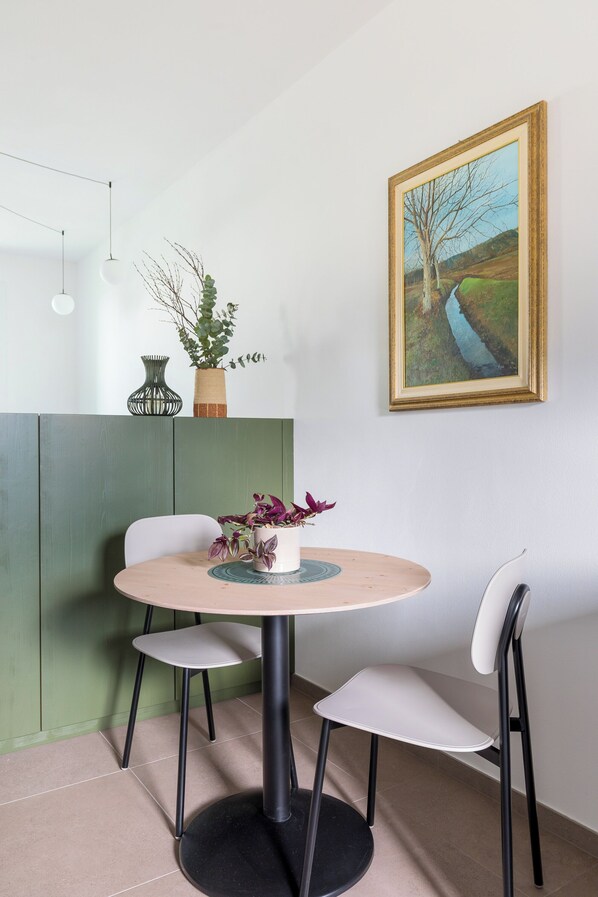 Ein Essbereich mit einem runden Tisch, zwei Stühlen, einer Blumenvase, einem Bild an der Wand und einem grünen Raumteiler.