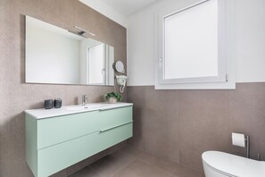 Ein modernes Badezimmer mit einem weißen Waschbecken, einer hellgrünen Kommode, einem großen Spiegel, einer Toilette und einem Fenster.