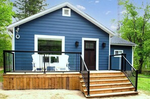 A blue house with a wooden deck, a dark brown door, and a small attached structure. - Nature's Playground - Three Bedroom Cottage, Sleeps 6 (Gravenhurst)