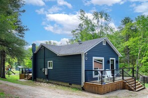 Une petite cabane bleu foncé, dotée d’une terrasse en bois et d’un porche abrité, entourée d’arbres.