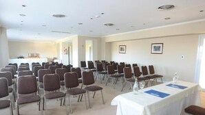 Une salle de conférence avec des rangées de chaises, une table recouverte d’une nappe blanche et, au fond, un coin cuisine.