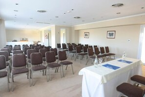 Un salón de conferencias con filas de sillas, una mesa con mantel blanco y una kitchenette al fondo.
