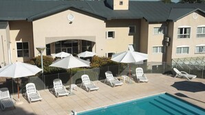 Un espace aménagé au bord de la piscine, avec des chaises longues et des parasols, une piscine à l’eau d’un bleu limpide, et un bâtiment à la façade beige ornée de nombreuses fenêtres.