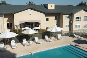 Zona de piscina con sillas de descanso y sombrillas, una piscina azul cristalina y un edificio con fachada color beige y numerosas ventanas.