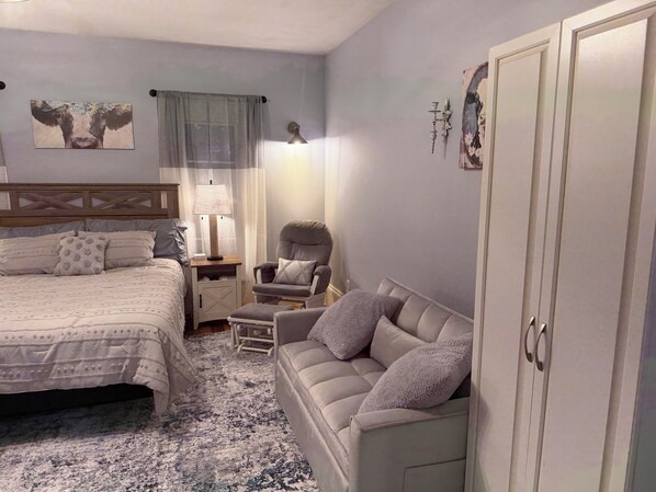 A bedroom with a bed, a sofa, a chair, a nightstand, a lamp, and a wardrobe.