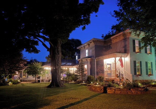 Una casa de dos pisos con un porche, una bandera y un jardín bien iluminado durante la noche.