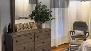Une chambre avec une commode en bois, un miroir, un panneau d’accueil, une plante en pot, une chaise et une lampe de chevet.