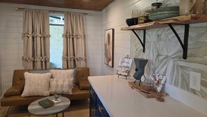 A cozy room with a wooden ceiling, a brown leather sofa with decorative pillows, a small round table with a book, and a kitchen area with a coffee maker and a shelf with kitchenware. - Tiny Blanco Retreat Birdhouse (Wimberley)