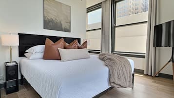 Een hotelkamer met een groot bed, een nachtkastje, een lamp, een schilderij aan de muur en uitzicht op gebouwen door het raam.