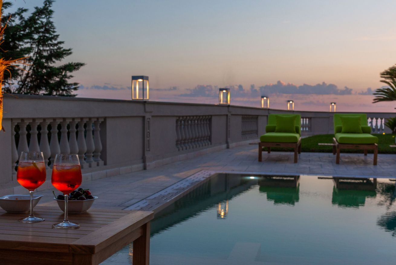 Zona de piscina al terrat amb dues cadira relaxants, una taula amb dues copes de beguda vermella, i vista al perfil urbà al vespre.