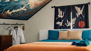 Ein Schlafzimmer mit einem großen Wandbild einer Welle, einem Bett mit türkisfarbenen und beigen Kissen, einer Holzkommode und einem hängenden Bademantel.