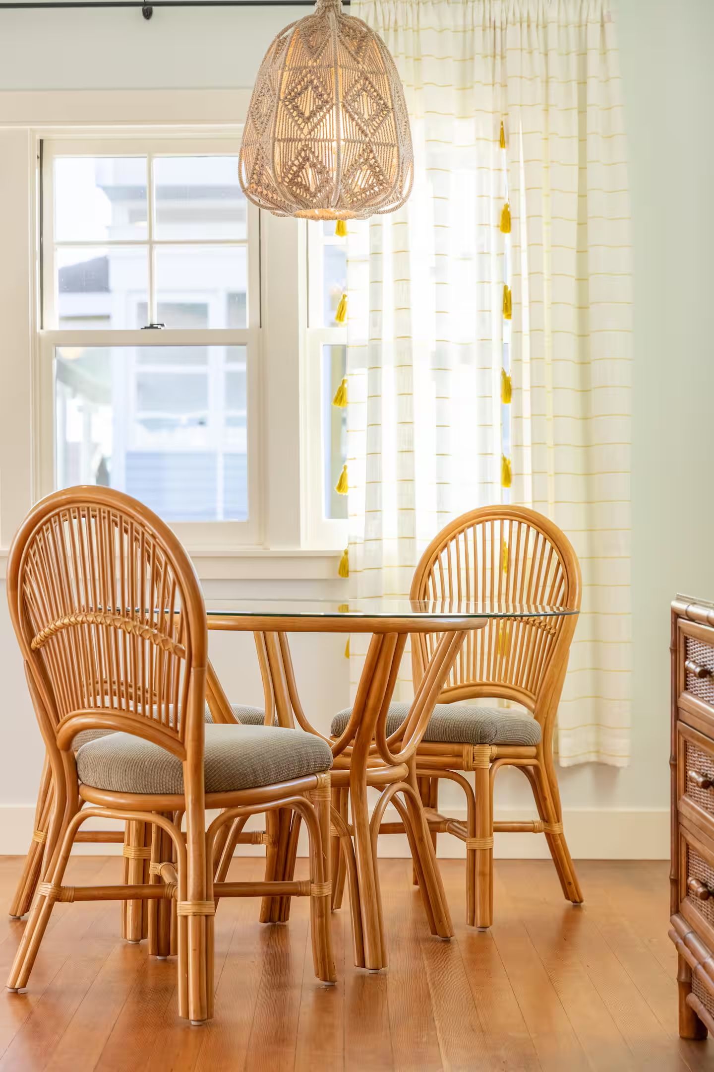 A dining area with a glass table and two wicker chairs, a hanging light fixture, and a window with curtains.