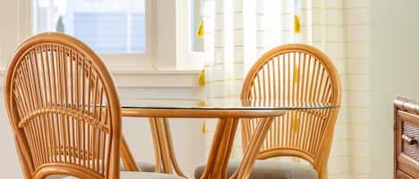 A dining area with a glass table and two wicker chairs, a hanging light fixture, and a window with curtains.