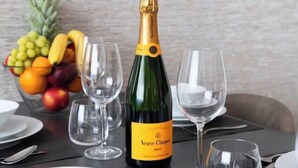 Ein Tisch, gedeckt mit einer Flasche Veuve Clicquot Champagner, leeren Weingläsern, weißen Tellern und einer Obstschale.