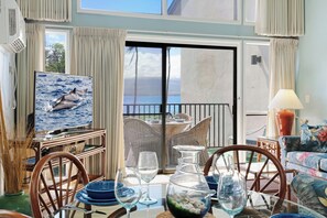 Une pièce dotée d’un téléviseur diffusant des images de dauphins, d’une table à manger avec des chaises, d’un canapé et d’une vue sur l’océan par le biais d’une porte coulissante.