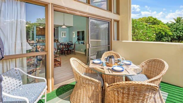 A balcony with wicker chairs and a glass table set for dining, a sliding glass door leading to an interior dining area, and a view of lush greenery. - Ocean-view 2BR Maui condo with pool, A/C, beach (Wailuku)