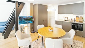 A modern living room with a round wooden dining table, white chairs, a painting on the wall, and a kitchen area with cabinets and a sink.