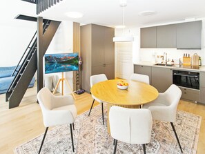 Una sala moderna con una mesa redonda de madera, sillas blancas, un cuadro en la pared y una zona de cocina con armarios y un lavamanos.