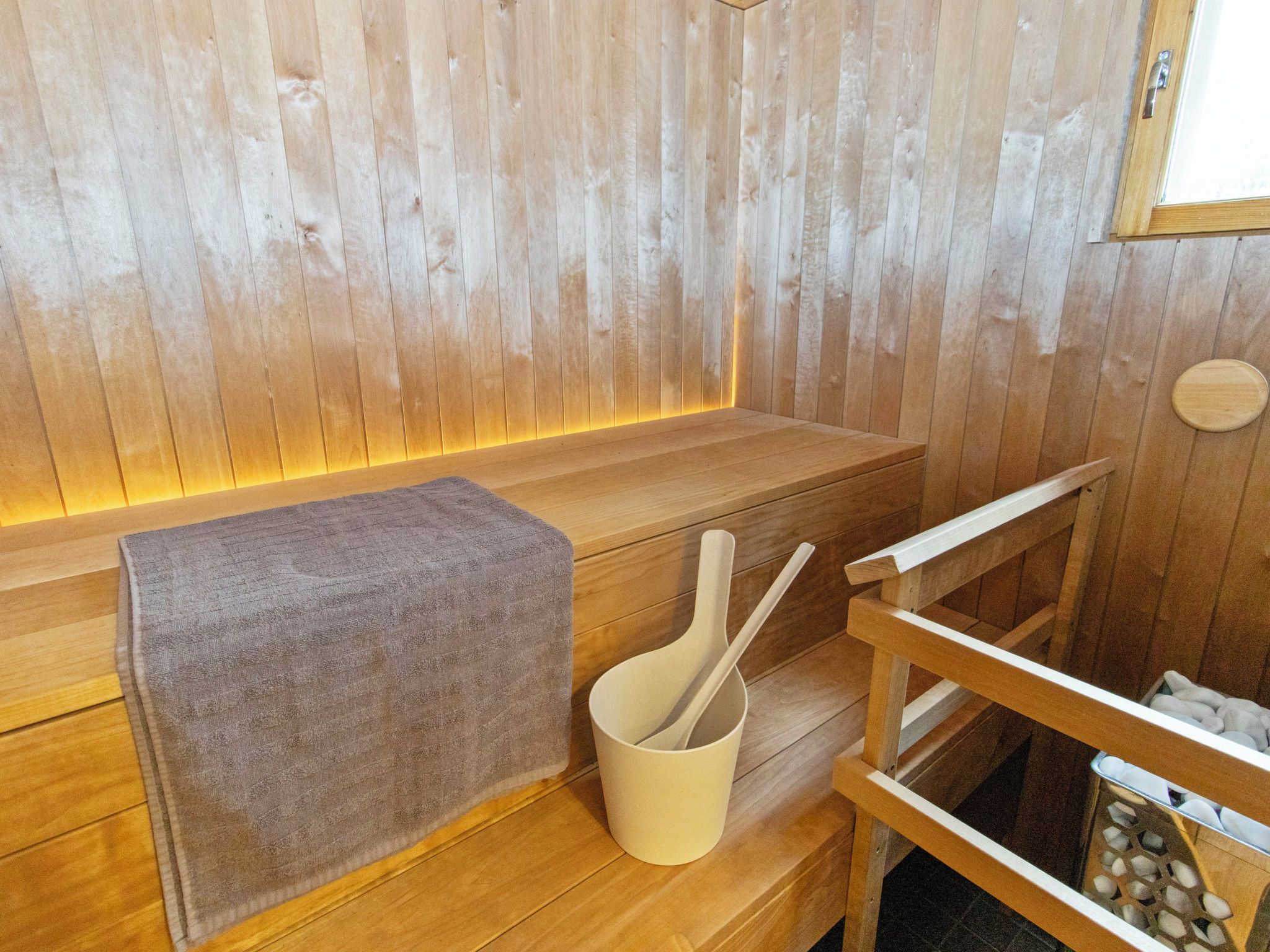 A wooden sauna room with a bench, a towel, and a wooden chair with a cup and spoon.