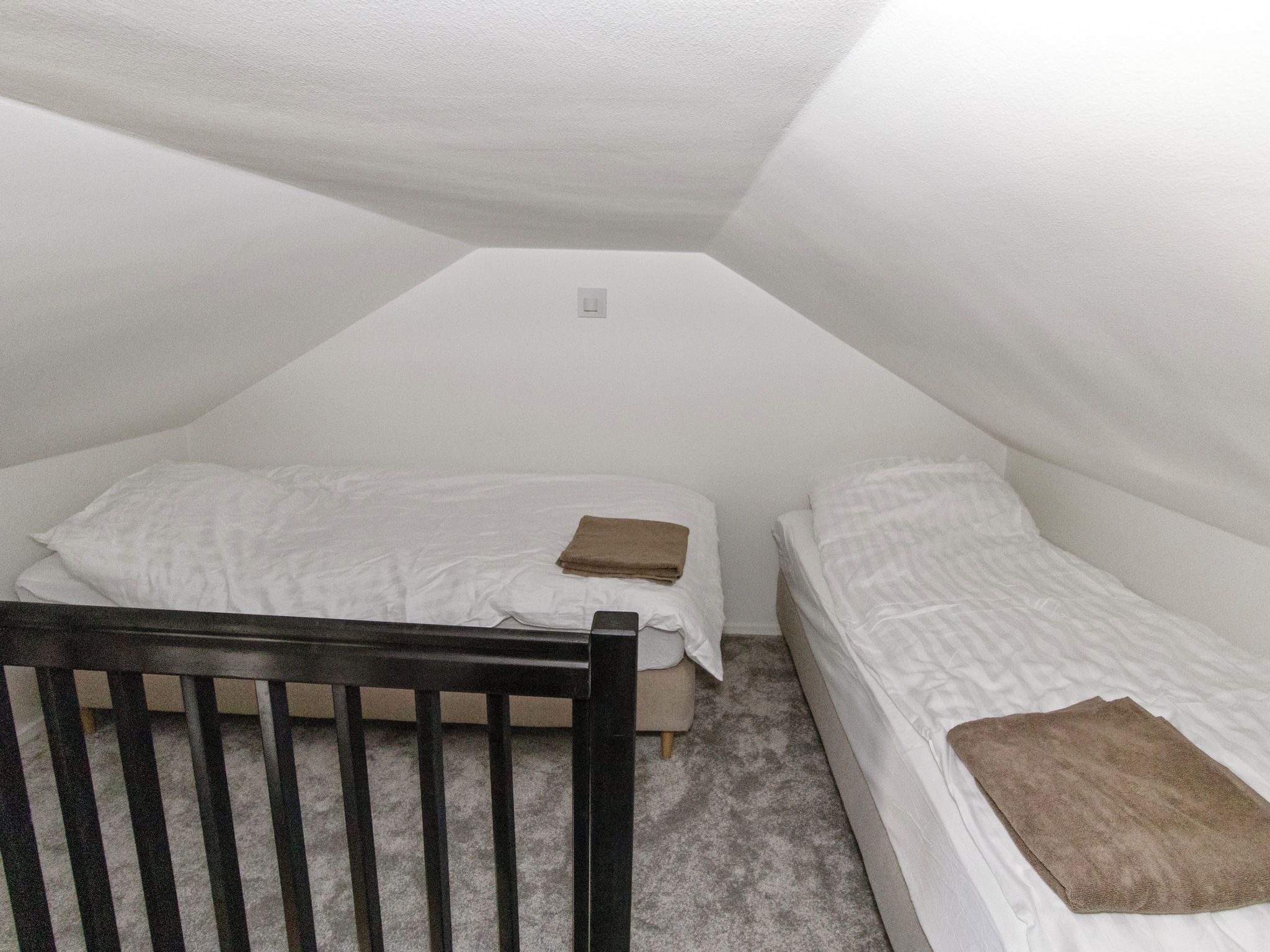 Een kleine kamer met een enkel bed, schuine wand, houten hoofdbord en een klein nachtkastje.