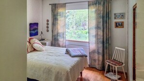 Ein Schlafzimmer mit Bett, Sessel, Schreibtisch mit Lampe, Fenster mit Vorhängen und Blick ins Grüne.