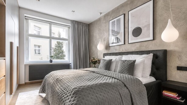 A modern bedroom with a large bed, a bedside table with books, a window with curtains, and wall art. - Stylish Apartment in Prague's Historic Centre (Prague 1)
