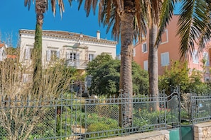 Ein Gebäude mit Balkon, umgeben von einem Garten mit Palmen und einem Metallzaun.