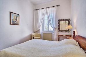 Ein Schlafzimmer mit Bett, Sessel, Schreibtisch mit Spiegel und Fenster mit Vorhängen.