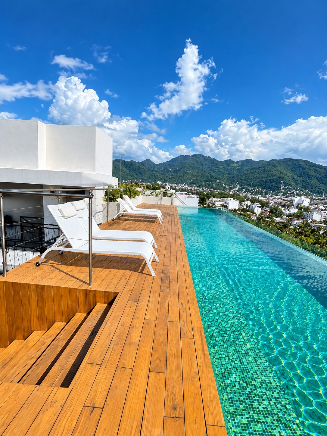 Une piscine à débordement située sur le toit, avec une terrasse en bois et des chaises longues blanches, offrant une vue imprenable sur un paysage montagneux.
