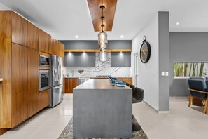A modern kitchen with wooden cabinets, stainless steel appliances, a marble island, and a dining area with blue chairs. - 4BR Family Villa with Pool, Gym & Private Parking (Nuevo Vallarta)