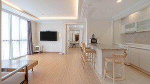Une chambre d’hôtel moderne dotée d’une télévision à écran plat, d’un coin repas avec des chaises en bois, d’une cuisine avec des meubles blancs et d’une grande fenêtre laissant entrer la lumière naturelle.
