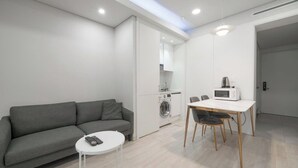 Un espace de vie moderne comprenant un canapé gris, une petite table blanche, une table à manger avec des chaises, un lave-linge et un four à micro-ondes.