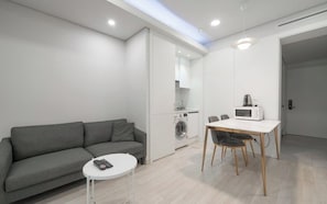 A modern living space with a grey sofa, a small white table, a dining table with chairs, a washing machine, and a microwave.