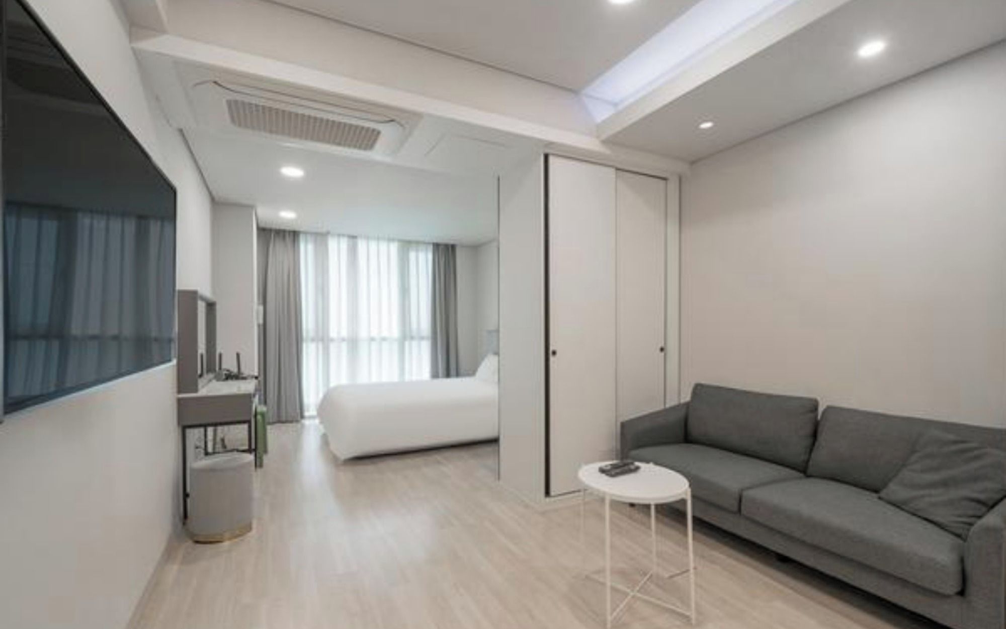 A modern hotel room with a grey sofa, a small round table, a bed, and a walk-in closet.