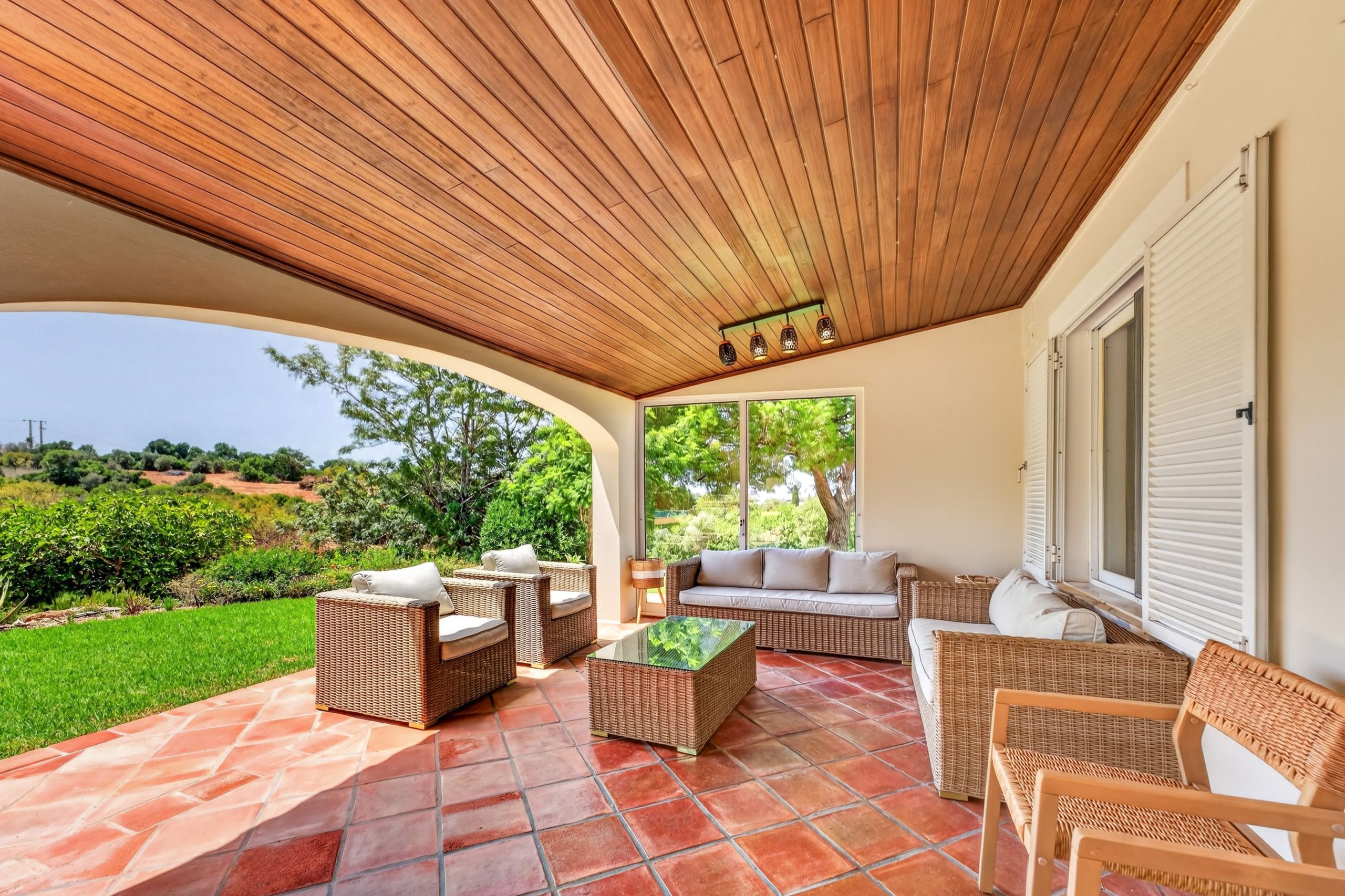A covered patio area with wicker furniture, a glass coffee table, and a view of the surrounding greenery.