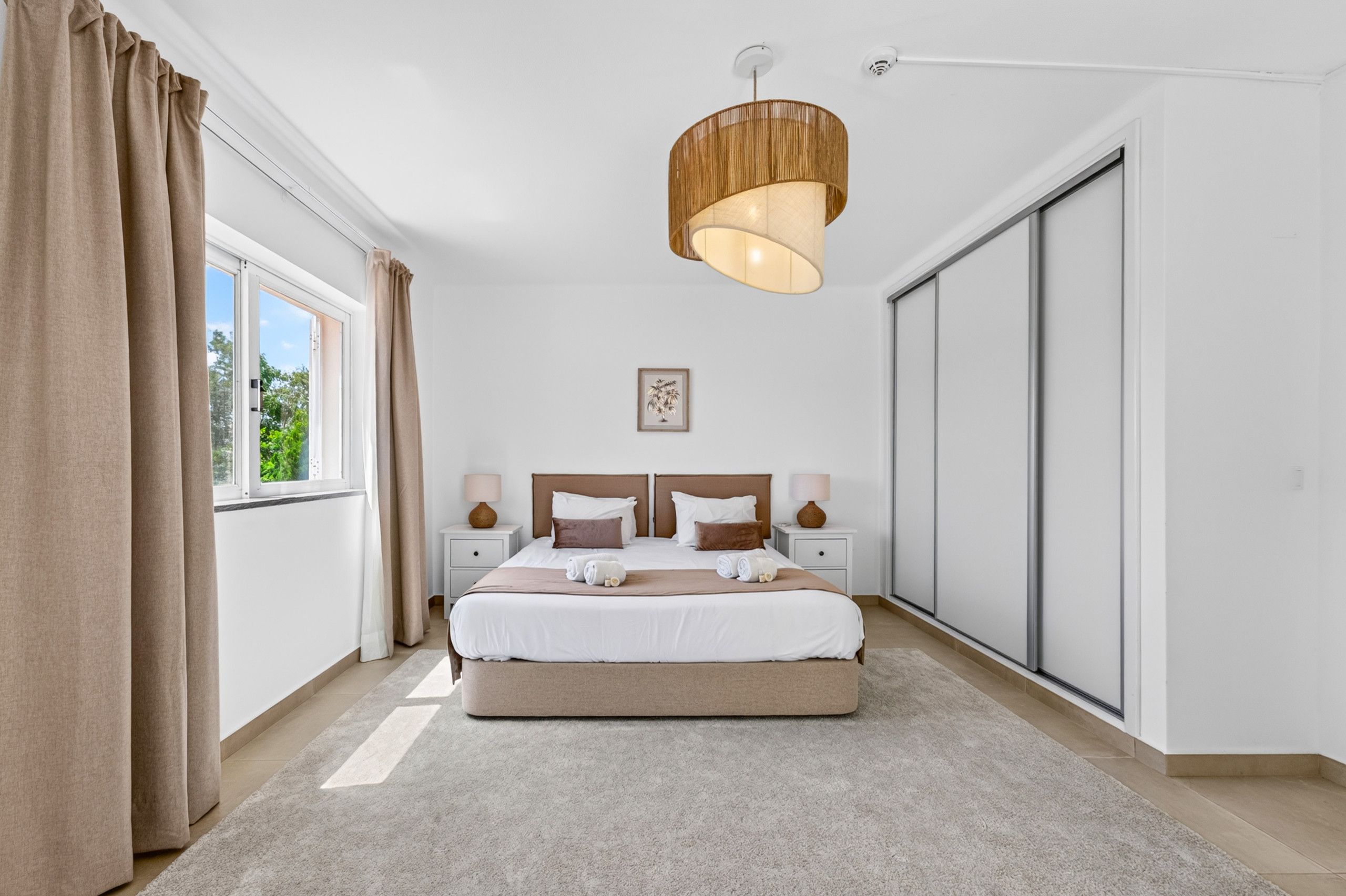A bedroom with a large bed, two bedside tables, a hanging light fixture, a window with curtains, and a sliding door.