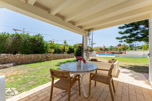 A covered patio with a round table and two wicker chairs, a stone wall, and a view of a swimming pool and greenery. - Villa Ursa | 6 bedrooms | Jacuzzi | Benagil (Carvoeiro)