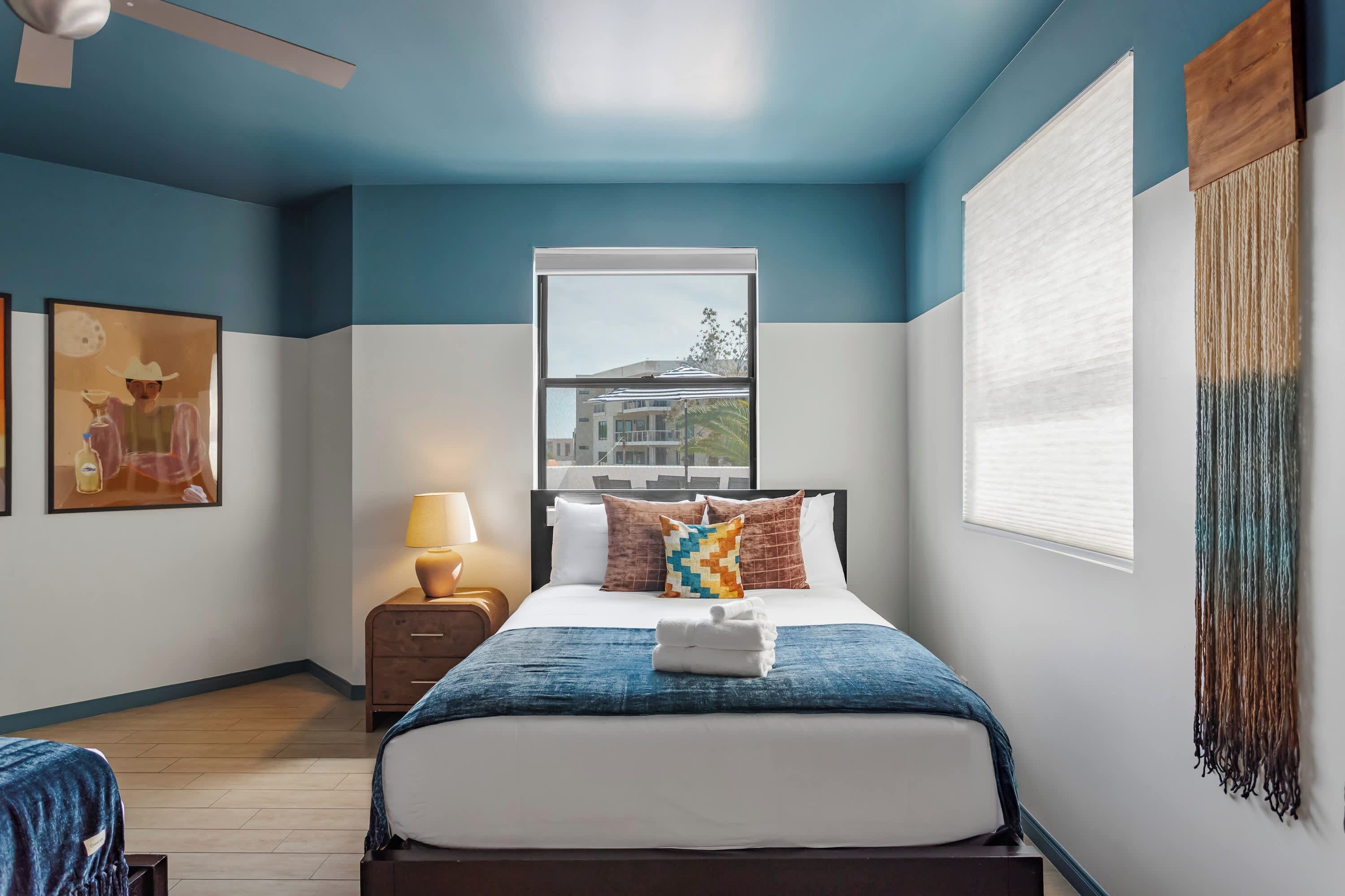 A hotel room with a bed, a nightstand, a lamp, a window, and a painting on the wall.
