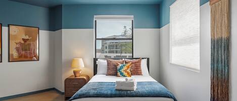 A hotel room with a bed, a nightstand, a lamp, a window, and a painting on the wall.