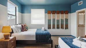 Une chambre d’hôtel avec un lit, un bureau, une chaise, une fenêtre, un ventilateur de plafond et une salle de bain visible à travers une porte ouverte.