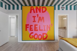 A room with a large wall mural, a bed with a yellow blanket, and a kitchen area visible through an open door. - Zeta - Private Balcony - In Old Town Scottsdale (Scottsdale)