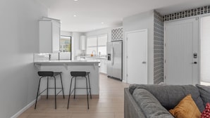Una cocina moderna con armarios blancos, una isla central con taburetes, un refrigerador y una zona de comedor con un sofá gris.