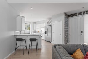 Eine moderne Küche mit weißen Schränken, einer zentralen Kochinsel mit Hockern, einem Kühlschrank und einem Essbereich mit grauem Sofa.
