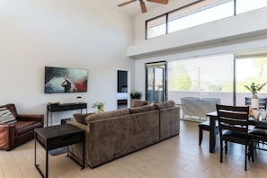 Ein modernes Wohnzimmer mit einer Couch, einem Couchtisch, einem Esstisch mit Stühlen, einem Deckenventilator und einem großen Fenster.
