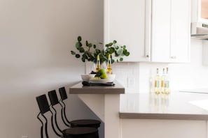 Eine moderne Küche mit weißen Schränken, einer Arbeitsplatte mit Obstschale und Flaschen sowie einem schwarzen Stuhl.